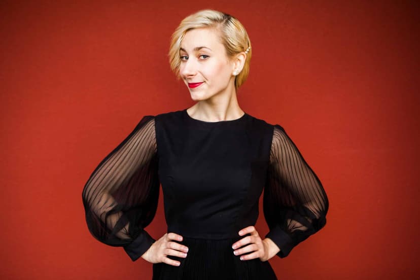 Natalia Eristavi in a black dress with sheer sleeves against a red wall